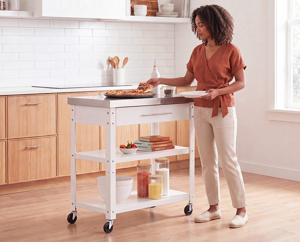 A woman with curly hair, wearing a terracotta wrap top and beige trousers, serves a slice of tart from a white rolling kitchen island. The cart features a stainless steel top, a drawer, and lower open shelves styled with cookware and cookbooks in a bright, modern kitchen.