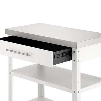 A close-up view shows the upper portion of a white kitchen cart with a stainless steel countertop against a plain background. Its single top drawer is pulled partially open on dark metal glides, revealing two open storage shelves positioned below.