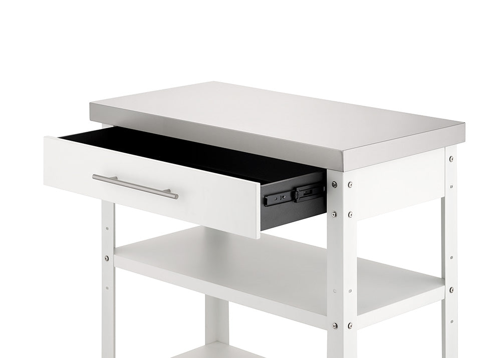 A close-up view shows the upper portion of a white kitchen cart with a stainless steel countertop against a plain background. Its single top drawer is pulled partially open on dark metal glides, revealing two open storage shelves positioned below.