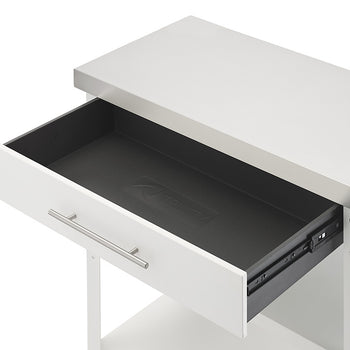 An angled close-up shot shows the top drawer of a white kitchen cart pulled open beneath a stainless steel countertop. The interior of the drawer is empty, featuring a black drawer liner embossed with the Trinity brand logo.