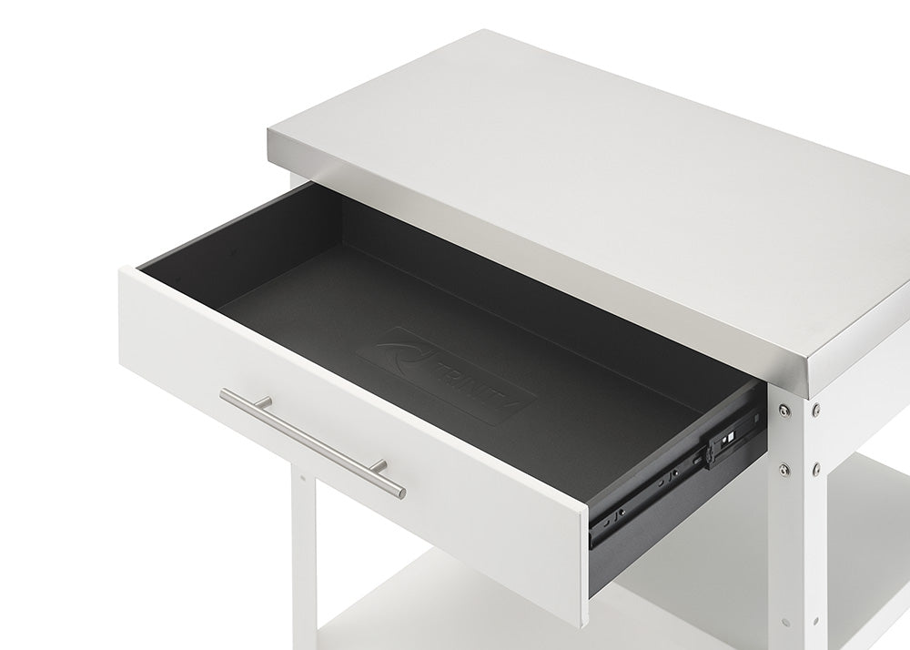 An angled close-up shot shows the top drawer of a white kitchen cart pulled open beneath a stainless steel countertop. The interior of the drawer is empty, featuring a black drawer liner embossed with the Trinity brand logo.