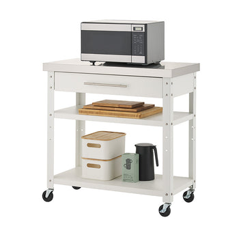 A white rolling kitchen cart featuring a stainless steel countertop and a single top drawer sits against a plain white background. A compact microwave is placed on the top surface, while the lower two shelves hold stacked wooden cutting boards, storage bins, a box, and a black pitcher.