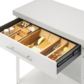 An angled close-up view shows the open drawer of a white kitchen cart topped with a stainless steel surface. Inside, customizable bamboo drawer organizers hold small dining accessories, including spoons, salt and pepper shakers, coasters, tongs, and a cake server.