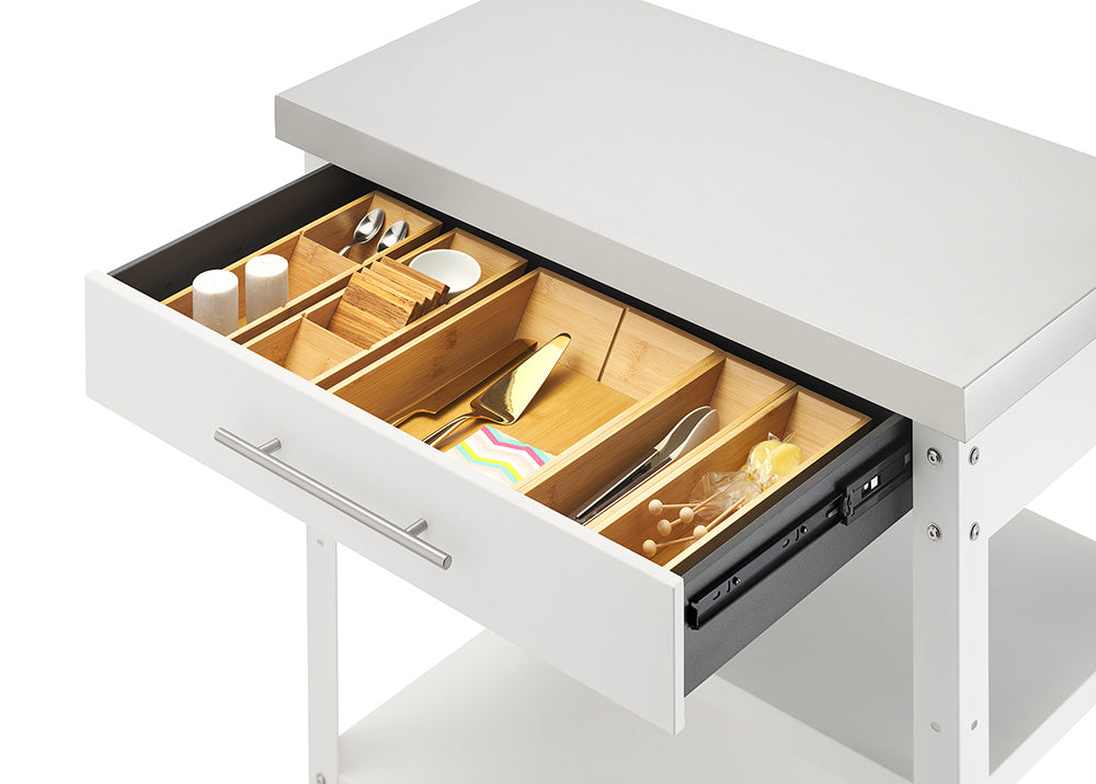 An angled close-up view shows the open drawer of a white kitchen cart topped with a stainless steel surface. Inside, customizable bamboo drawer organizers hold small dining accessories, including spoons, salt and pepper shakers, coasters, tongs, and a cake server.