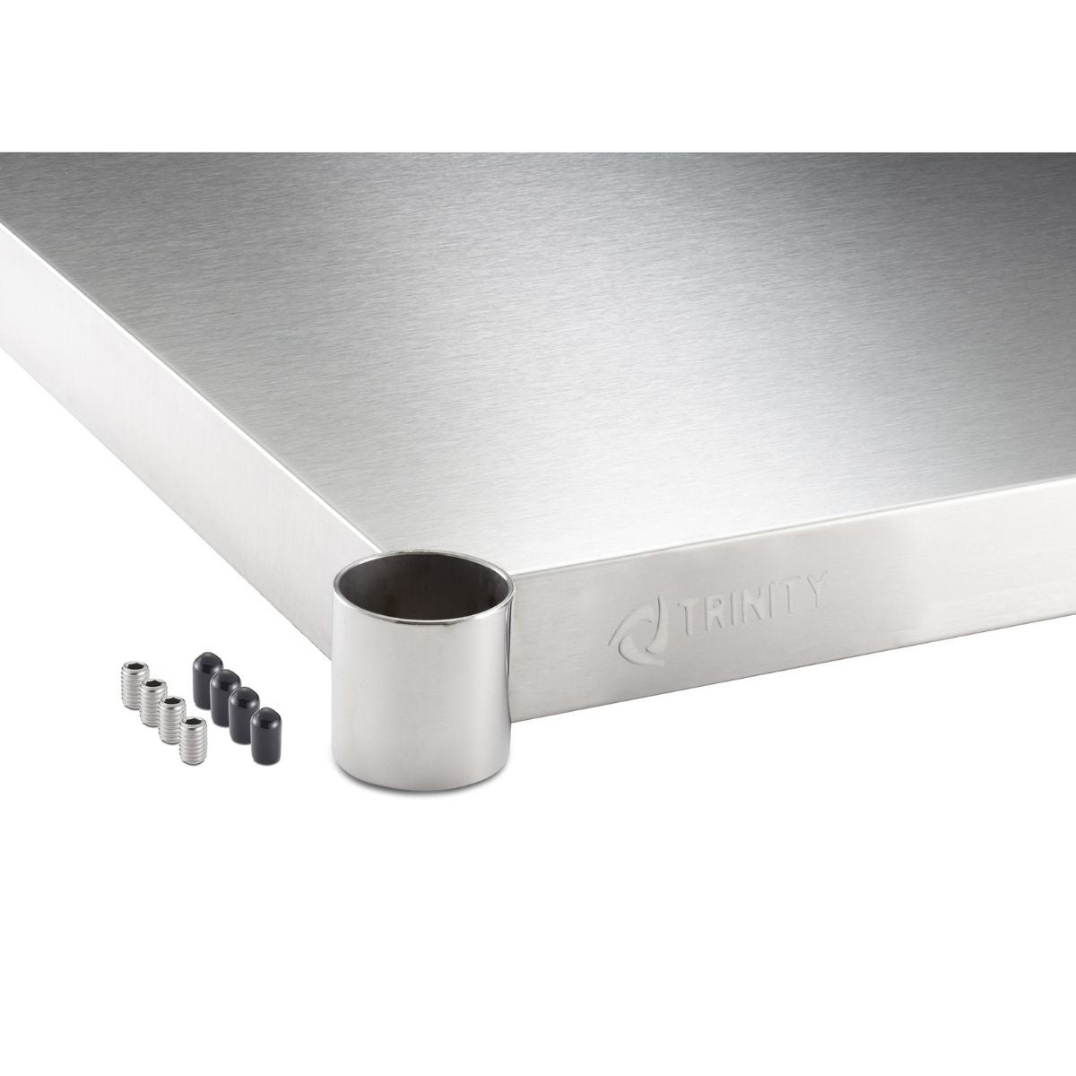 A close-up shows the corner of a stainless steel work surface with the word "TRINITY" subtly embossed on the side. Next to the corner are a cylindrical stainless steel leg piece, four small black caps, and four small silver screws.