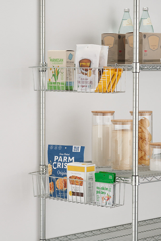 A close-up shot shows two wire metal hanging baskets attached to a chrome shelving unit. The baskets hold various pantry items, including boxes of crackers, a bag of coffee, a container of parmesan cheese, and jars of dry goods.