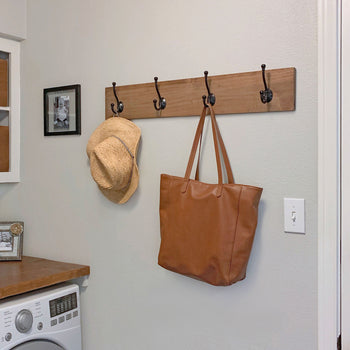 A wooden wall-mounted coat rack with five metal hooks holds a straw hat and a brown leather tote bag. The rack is installed in a laundry or mudroom with a washing machine, framed photos, and wooden shelves nearby.
