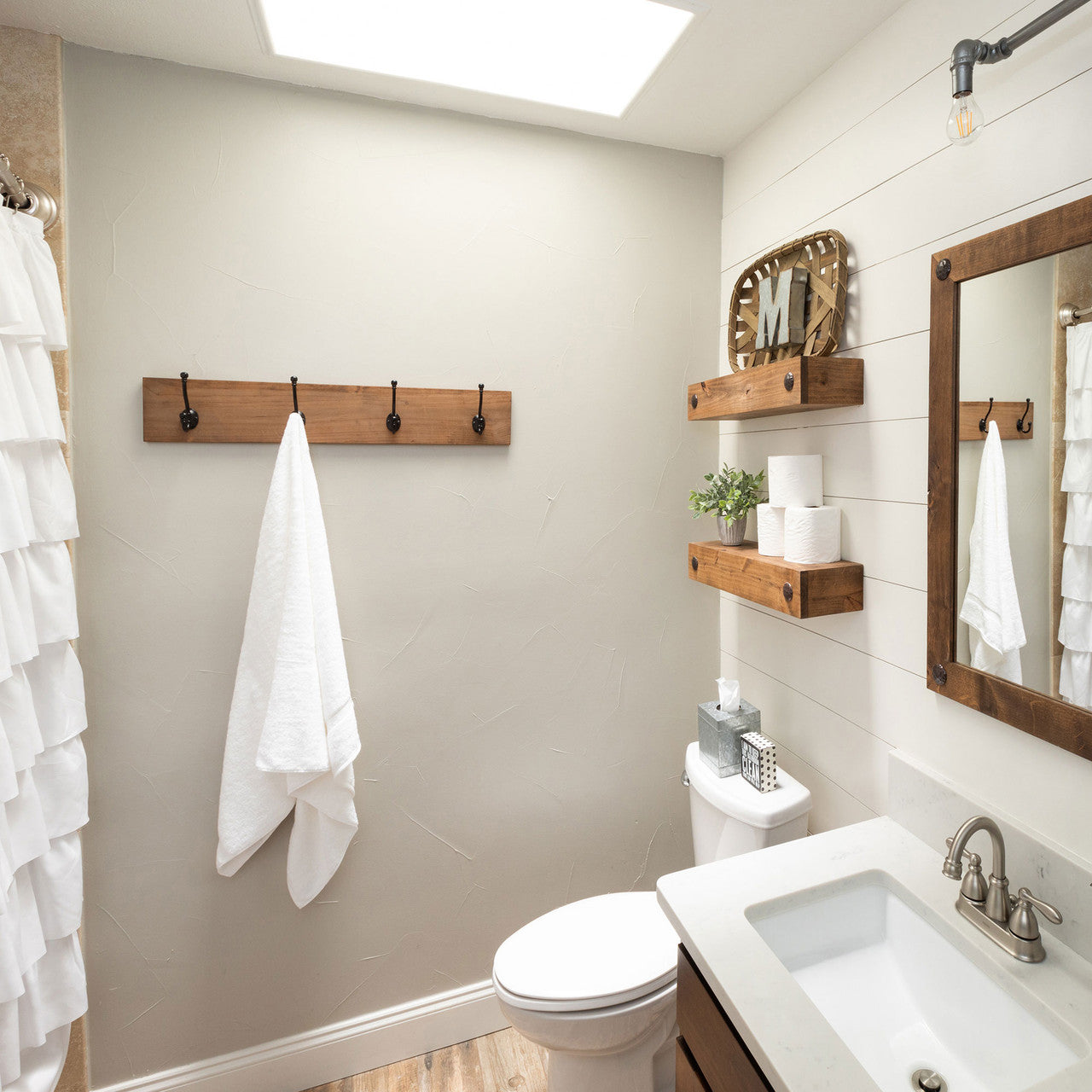 A modern farmhouse-style bathroom features wooden accents, a white sink, and a toilet. The space is decorated with floating wooden shelves, a wooden-framed mirror, and a towel hanging on a rustic wooden hook rack.