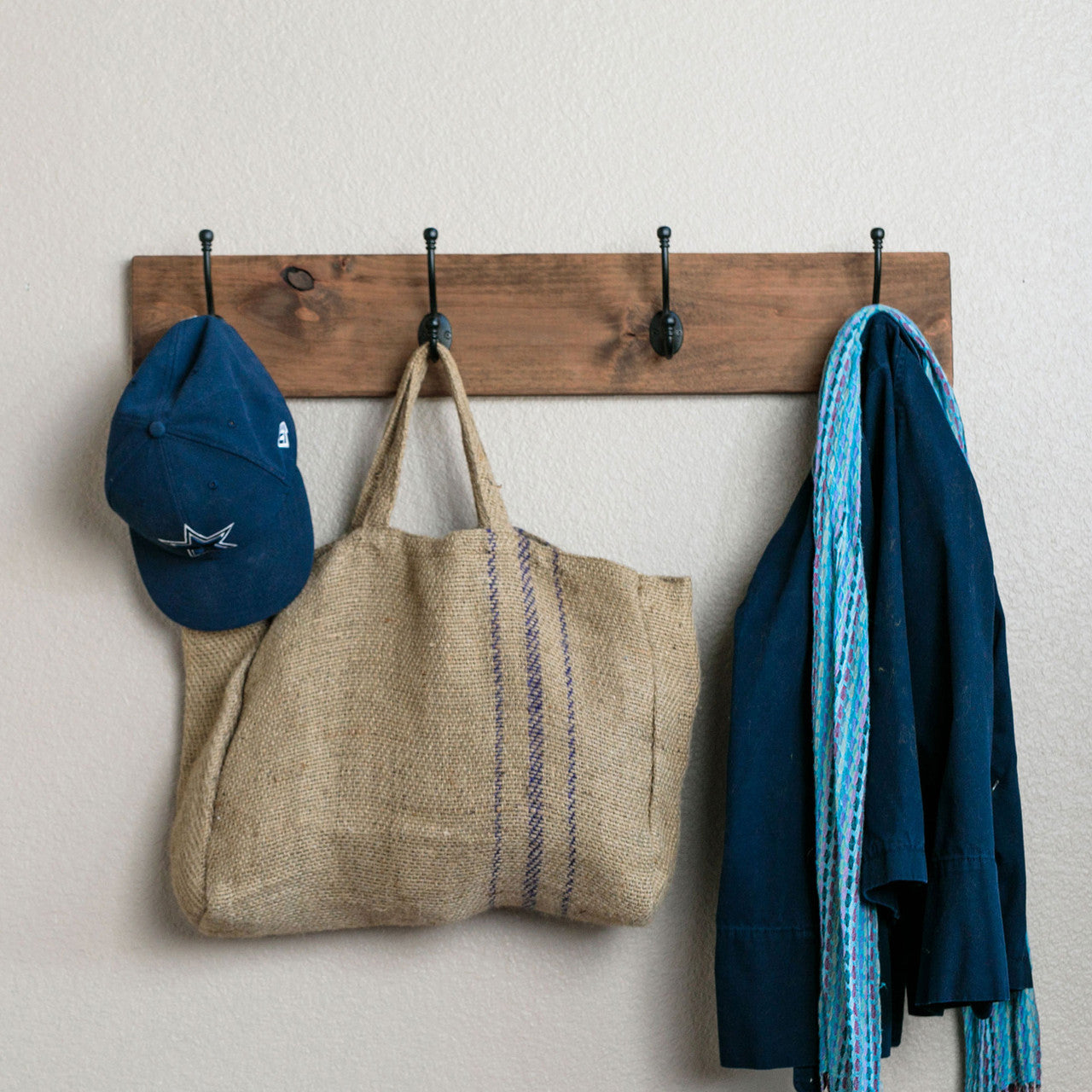 A wooden wall-mounted coat rack with four black metal hooks holds a navy blue baseball cap, a burlap tote bag with purple stripes, a dark blue jacket, and a blue patterned scarf. The rack is fixed to a light beige wall, and the items hang neatly in an organized manner.
