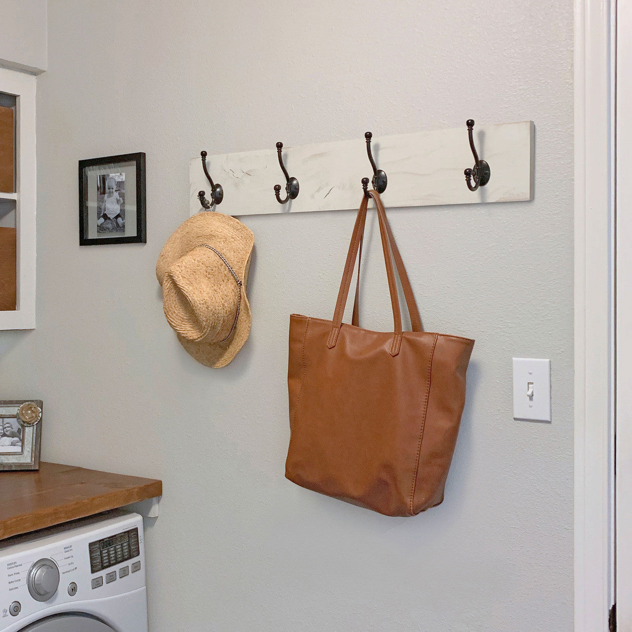 A whitewashed wooden wall-mounted coat rack with five black metal hooks is attached to a light gray wall. A brown leather tote bag and a straw hat hang from two of the hooks, while a washing machine and wooden countertop are visible below.