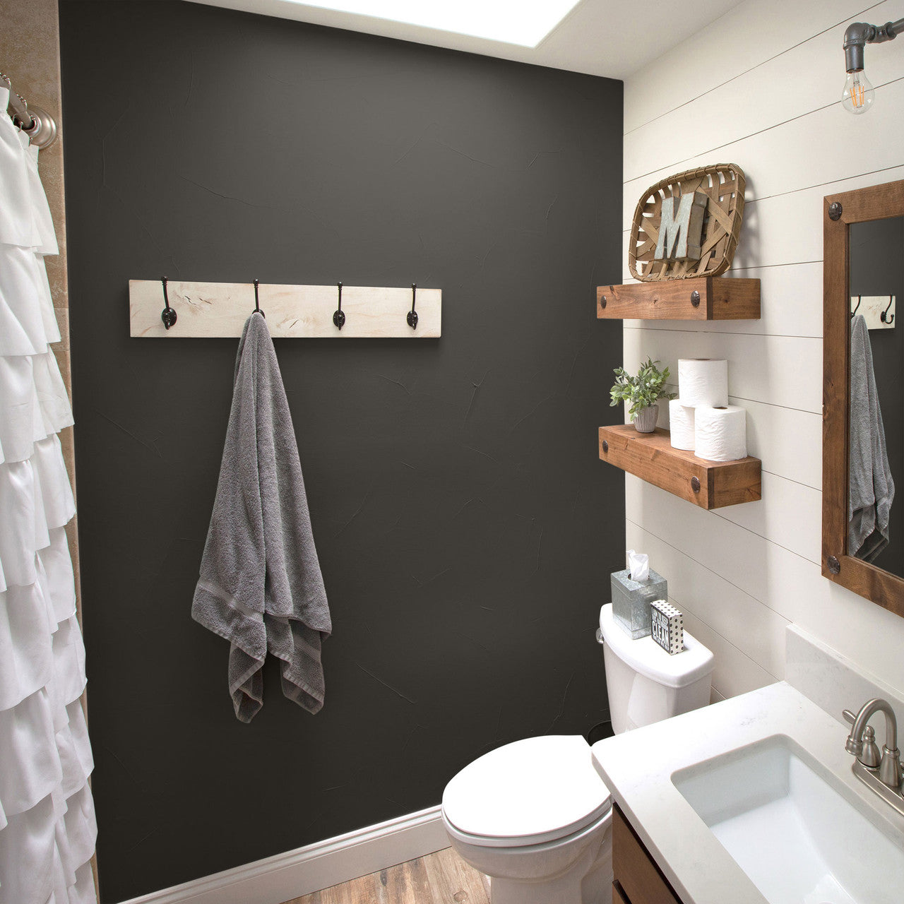 A modern farmhouse-style bathroom features a dark accent wall with a wooden towel rack holding a gray towel. The space includes rustic wooden shelves, a white vanity, and a toilet, decorated with small plants and neatly stacked toilet paper rolls.